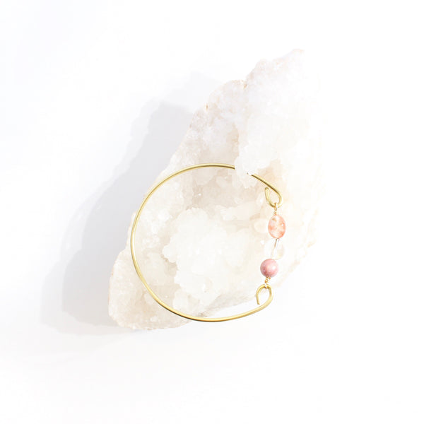 gold wire bangle with two sides held together by rhodonite, clear quartz and sunstone beads. top view, laying on a bed of white crystals. white background