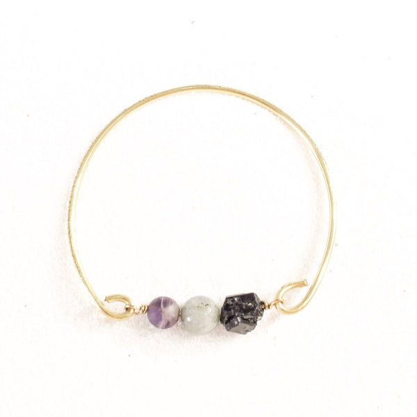 white background with a gold brass bangle. on the bangle is raw back tourmaline, a labradorite sphere bead and an amethyst sphere bead. birds eye view.