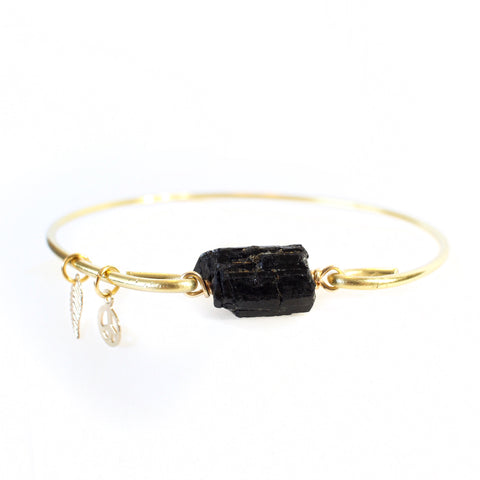 gold bangle with a chunk of black tourmaline in the middle, on the bangle are little gold charms, one is a little leaf and the other is a peace sign. it is sitting on a white background.