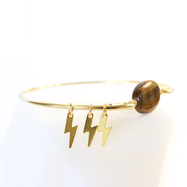 gold coloured bangle with tigers eye bead connecting the two sides. there are three little gold lightning bolt charms on the bangle. 45 degree view, close up of lightning bolt charms, white background