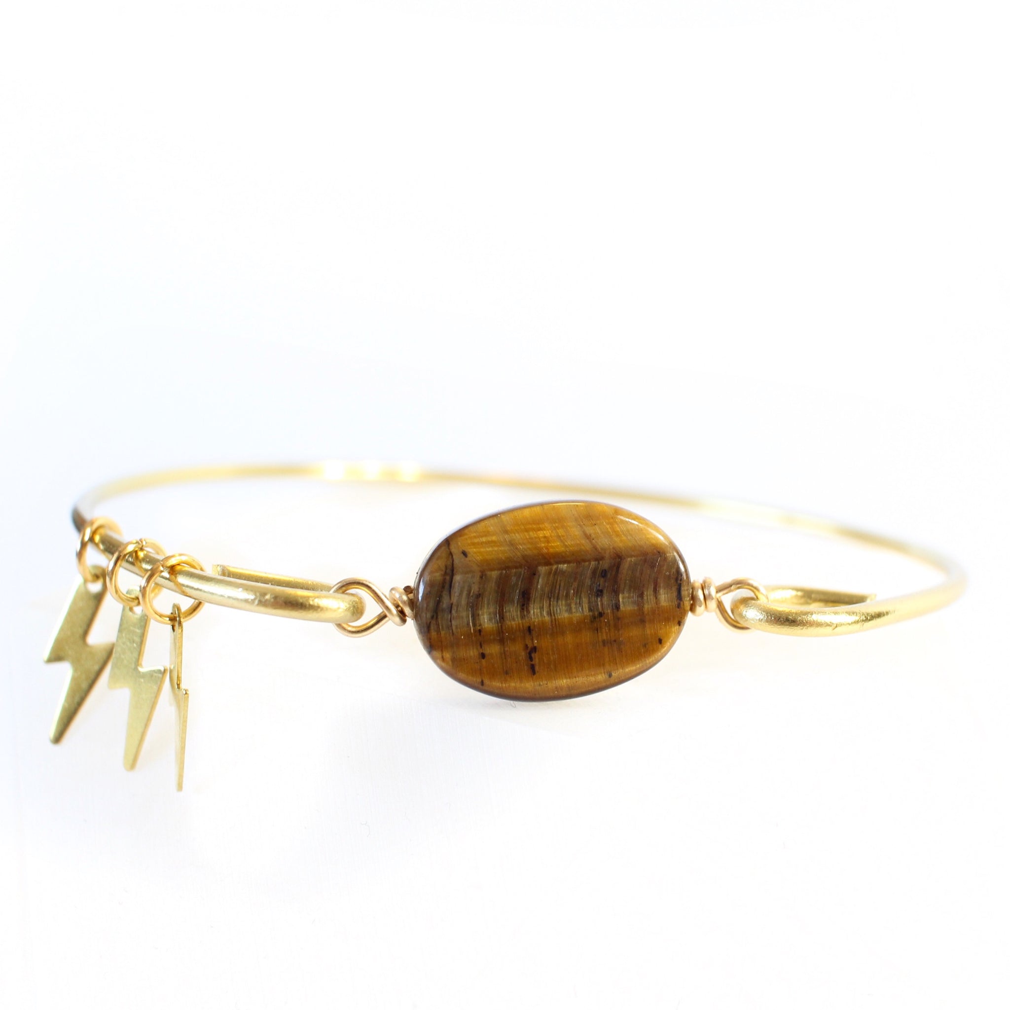 gold coloured bangle with tigers eye bead connecting the two sides. there are three little gold lightning bolt charms on the bangle. front close up view, white background