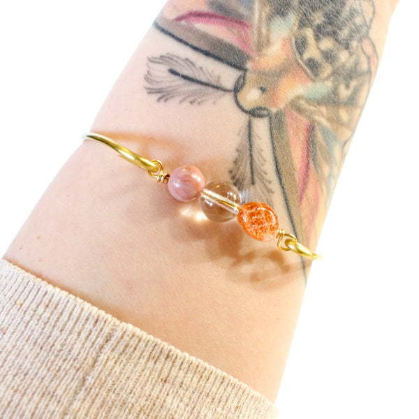 gold wire bangle with two sides held together by rhodonite, clear quartz and sunstone beads. shown worn on a tattooed wrist, white background