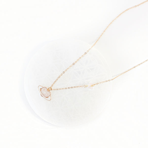 dainty gold chain necklace with a piece of rose quartz on the chain, there is an enamel pendant, a light pink heart with a white ring like a planet. necklace is laying on a selenite plate, with a white background