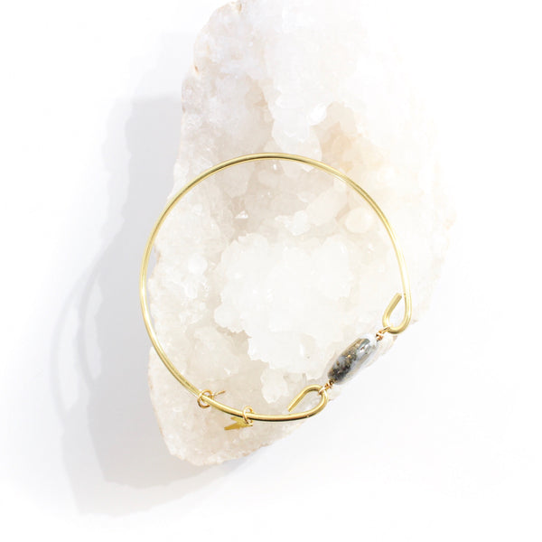 gold wire bangle with two sides held together with a pyrite and quartz coin shaped bead. there are little gold charms on the bangle, a star and a lightning bolt. the pyrite on the bead resembles the marbling on a planet. top view, shown resting on a bed of white crystals, white background.