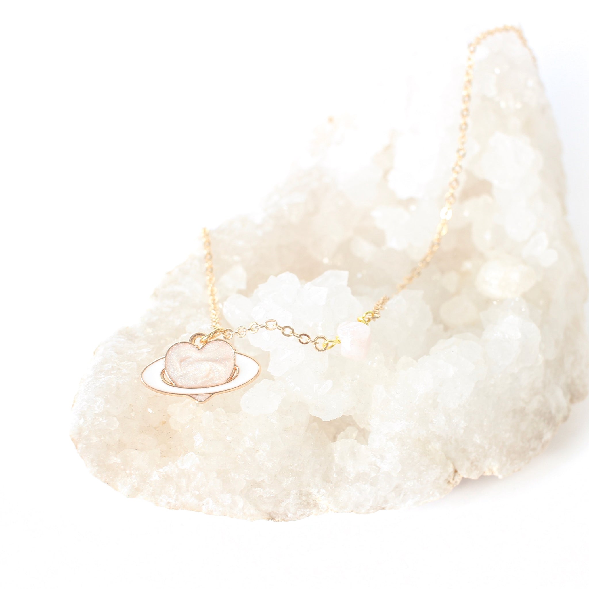 dainty gold chain necklace with a piece of rose quartz on the chain, there is an enamel pendant, a light pink heart with a white ring like a planet. necklace is laying on a quartz cluster bed, with a white background