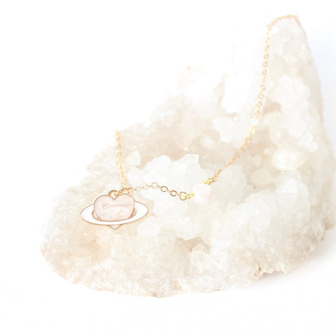 dainty gold chain necklace with a piece of rose quartz on the chain, there is an enamel pendant, a light pink heart with a white ring like a planet. necklace is laying on a quartz cluster bed, with a white background