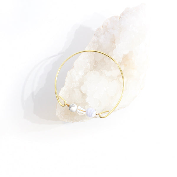 gold bangle with blue lace agate, howlite and quartz beads laying on crystal bed with white background