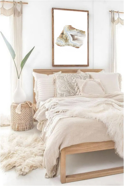 high quality photo of a blue lace agate crystal with a white background in a wood frame.  frame is hung on a white wall between two sheer curtains and above a natural wood bohemian bed with textured white pillows and blankets. beside the bed there is a table that looks like a woven basket with a simple tall plant on top