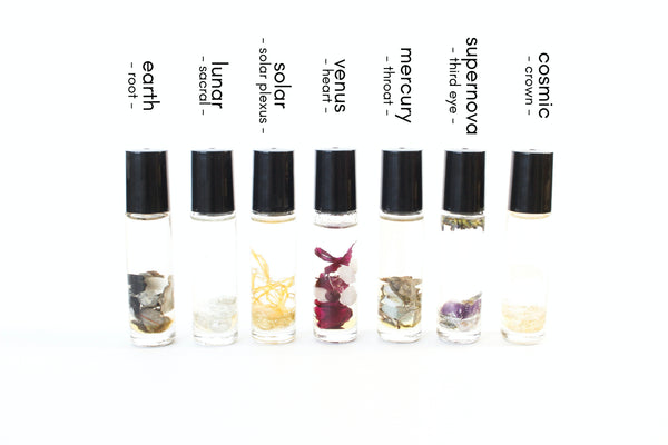 white background with 7 clear glass roller bottles with black lids. the bottles have holographic Gem+Aura logos. in the bottles there is an array of different crystals, dried flowers + herbs. There is one roller for each chakra- displaying the entire roller collection. 