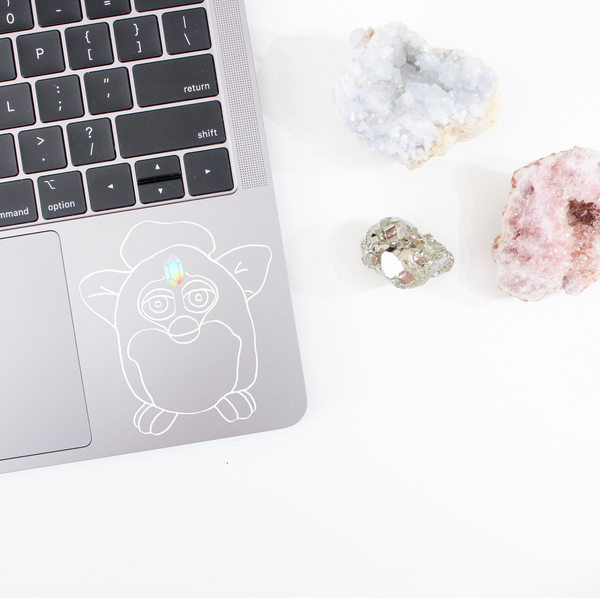 white decal mockup of a Furby on the bottom corner of a grey MacBook. instead of the Furby's sensor on it's forehead, there is a holographic crystal