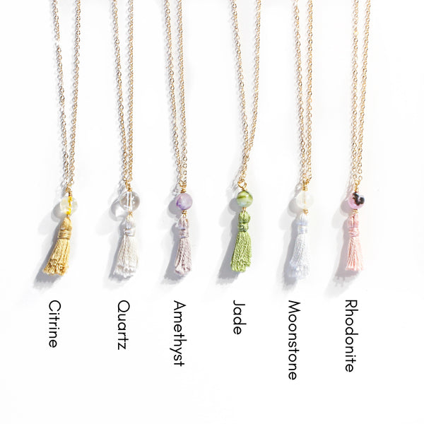 six necklaces, all with dainty gold chains, a crystal and a mini cotton tassel at the end. there is moonstone, citrine, rhodonite, amethyst, quartz and Jade. they are laying on a white background