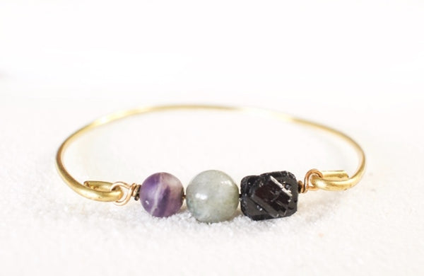 white background with a gold brass bangle. on the bangle is raw back tourmaline, a labradorite sphere bead and an amethyst sphere bead. resting on white sand.