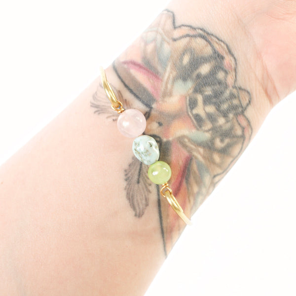 tattooed wrist wearing gold brass bangle with jade, larimar and rose quartz beads in the center. white background