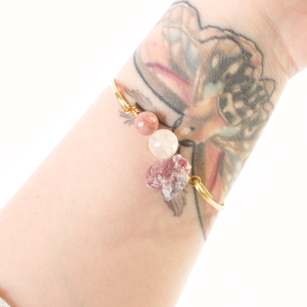 gold bangle with rhodonite, rose quartz and pink tourmaline beads in the middle. bracelet is shown modelled on a caucasian tattooed wrist and there is a white background