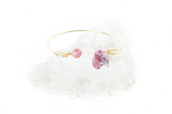 gold bangle with rhodonite, rose quartz and pink tourmaline beads in the middle. bracelet is resting on a bed of crystals and there is a white background