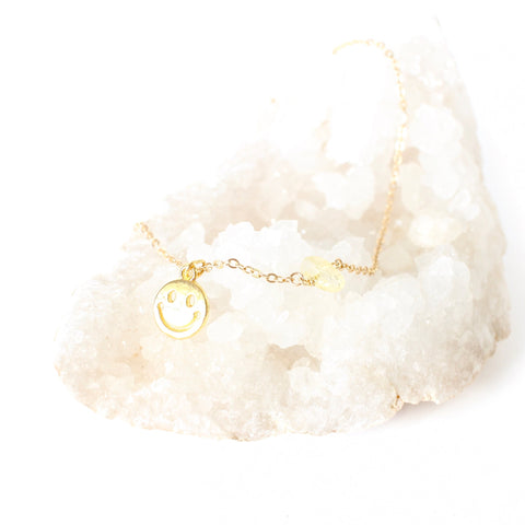 gold smiley face pendant on a dainty gold chain with a little piece of citrine on the chain. necklace is laying on a bed of white crystals with a white background