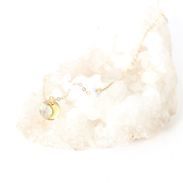 dainty gold necklace with a little moon pendant layered on top of a holographic looking shell pendant, necklace is draped over a cluster of white crystals and there is a white background. 