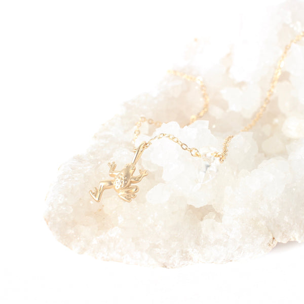 matte gold frog pendant on a dainty gold chain with a little piece of quartz on it. the necklace is laying on  cluster of white crystals, with a white background.