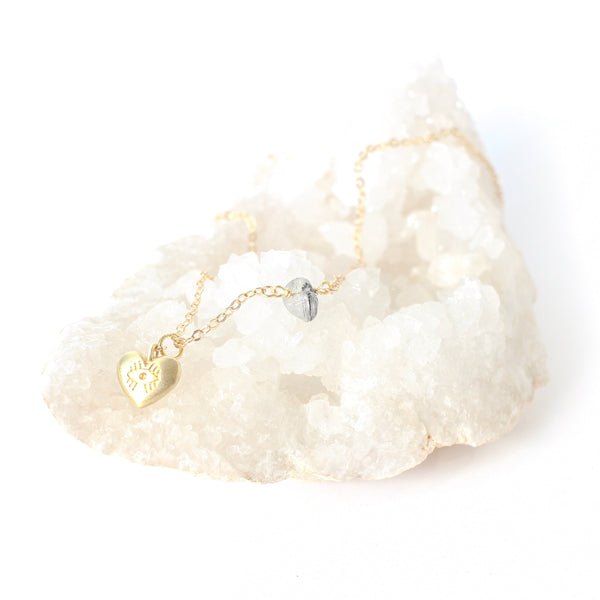 Matte gold heart pendant with an eye carved into it; the pendant is on a dainty gold chain with a piece of tourmalinated quartz on it. the necklace is sitting on a bed of quartz and there is a white background