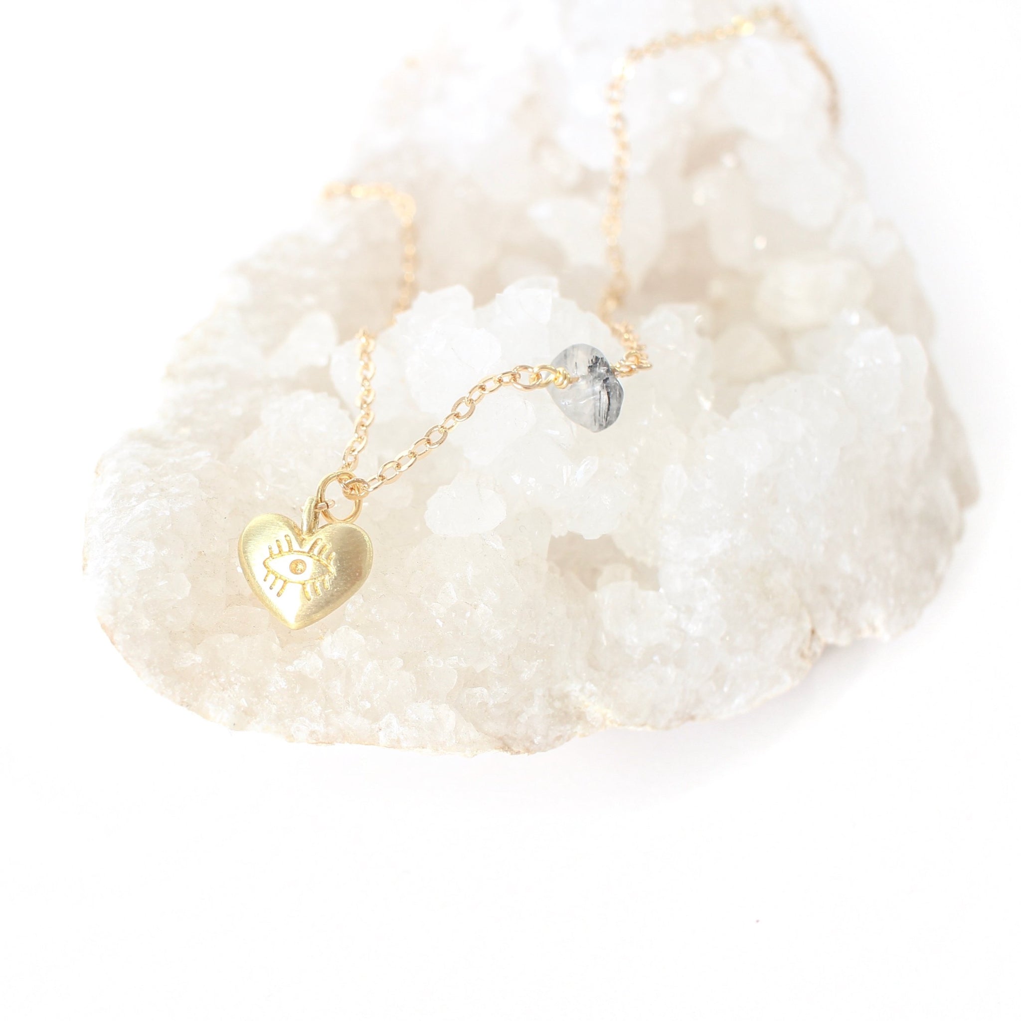 Matte gold heart pendant with an eye carved into it; the pendant is on a dainty gold chain with a piece of tourmalinated quartz on it. the necklace is sitting on a bed of quartz  and there is a white background