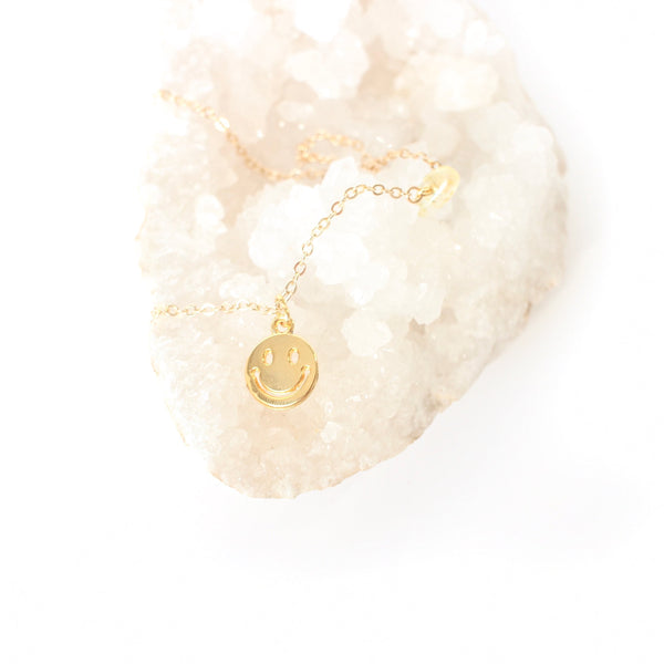 gold smiley face pendant on a dainty gold chain with a little piece of citrine on the chain. necklace is laying on a bed of white crystals with a white background