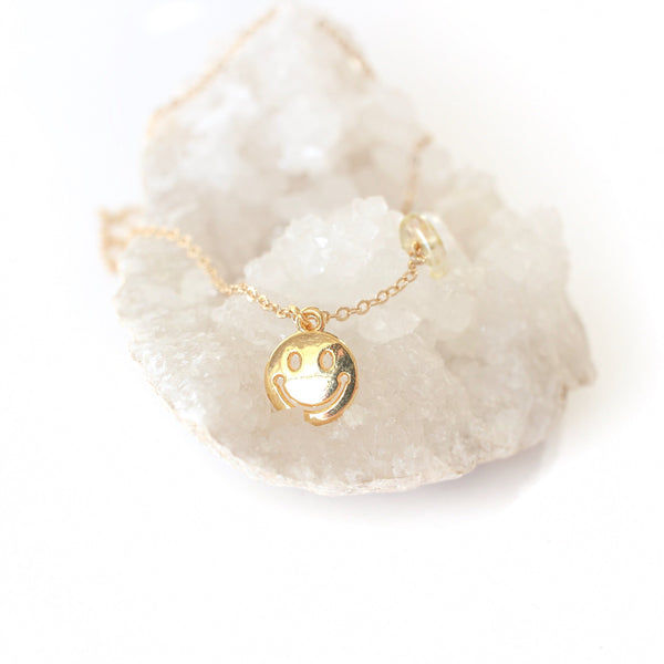 gold smiley face pendant on a dainty gold chain with a little piece of citrine on the chain. necklace is laying on a bed of white crystals with a white background