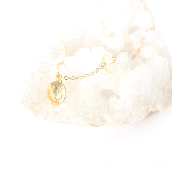 gold smiley face pendant on a dainty gold chain with a little piece of citrine on the chain. necklace is laying on a bed of white crystals with a white background