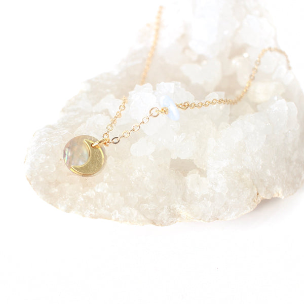 dainty gold necklace with a little moon pendant layered on top of a holographic looking shell pendant, necklace is draped over a cluster of white crystals and there is a white background. 