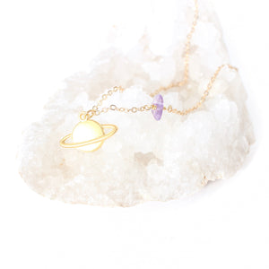 matte gold Saturn pendant on a dainty gold chain with a small amethyst piece on it. the necklace is laying on a white bed of quartz and there is a white background