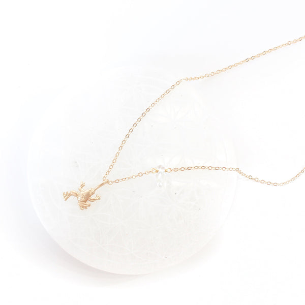 matte gold frog pendant on a dainty gold chain with a little piece of quartz on it. the necklace is laying on a white round selenite plate, with a white background.