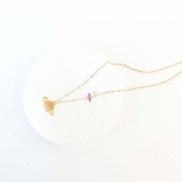 matte gold Saturn pendant on a dainty gold chain with a small amethyst piece on it. the necklace is laying on a white selenite plate and there is a white background