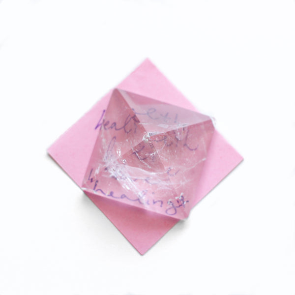 1.5 - 2 inch wide clear quartz pyramid, sitting on a pink piece of paper with writing underneath. photo is taken from top view, with a white background