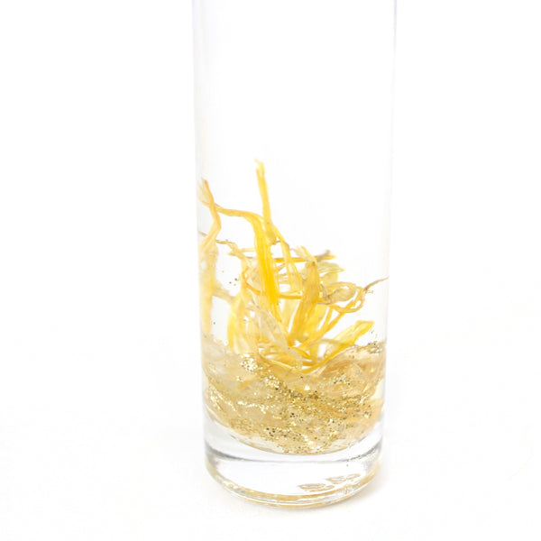 clear glass roller bottle. in the bottle there is an essential oil blend, and dried calendula, gold glitter and citrine chips. there is a white background. photo is a close up of the contents