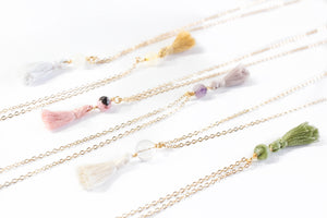 six necklaces, all with dainty gold chains, a crystal and a mini cotton tassel at the end. there is moonstone, citrine, rhodonite, amethyst, quartz and Jade. they are laying diagonally across the photo on a white background