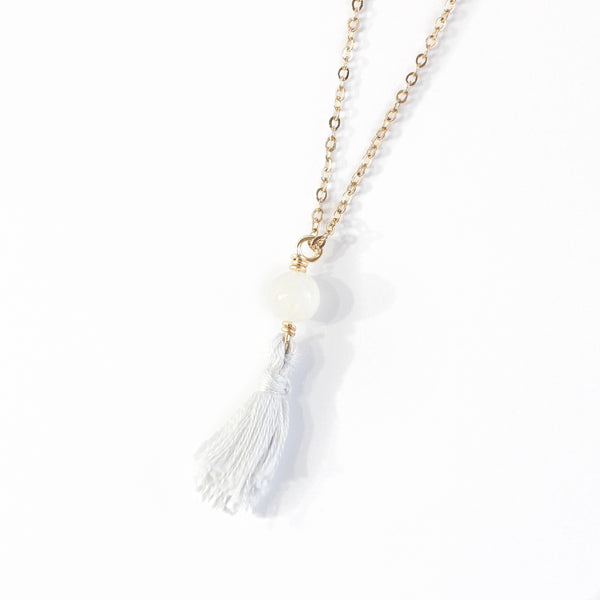 necklace with a  dainty gold chain, a moonstone bead and a light grey-blue mini cotton tassel at the end.. it is laying on a white background