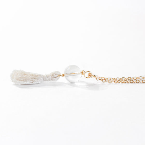 necklace with a dainty gold chain, a clear quartz bead and a cream mini cotton tassel at the end.. it is laying on a white background