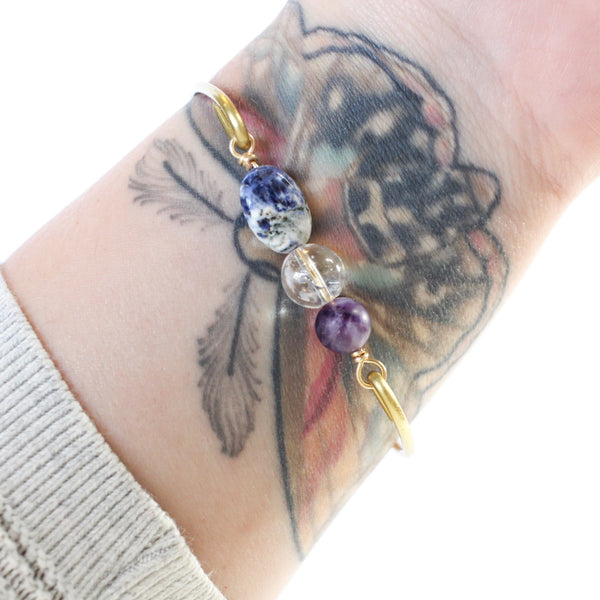 gold coloured bangle with amethyst, clear quartz and sodalite beads connecting the two sides of the wire bangle. white background, shown worn on a tattooed wrist