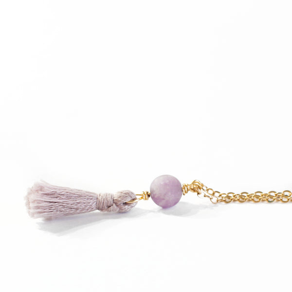 necklace with a dainty gold chain, an amethyst bead and a light mauve mini cotton tassel at the end.. it is laying on a white background