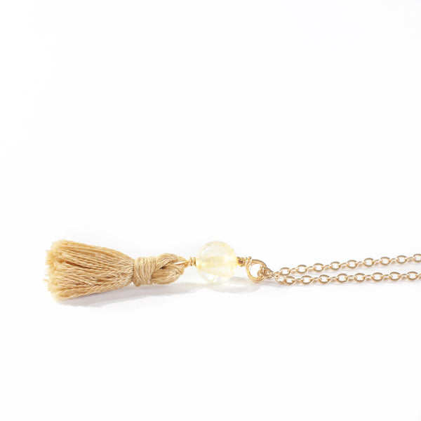 necklace with a dainty gold chain, a citrine bead and a camel/gold mini cotton tassel at the end.. it is laying on a white background