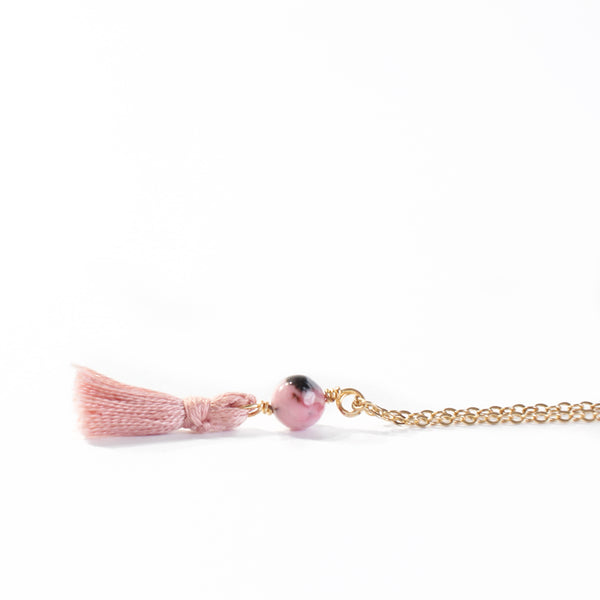 necklace with a dainty gold chain, a rhodonite bead and a dusty pink mini cotton tassel at the end.. it is laying on a white background