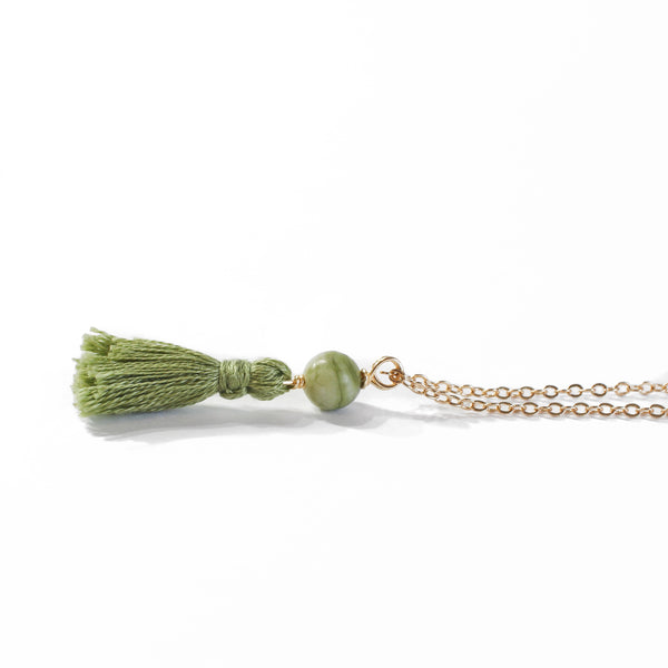 necklace with a dainty gold chain, a jade bead and an olive green mini cotton tassel at the end.. it is laying on a white background