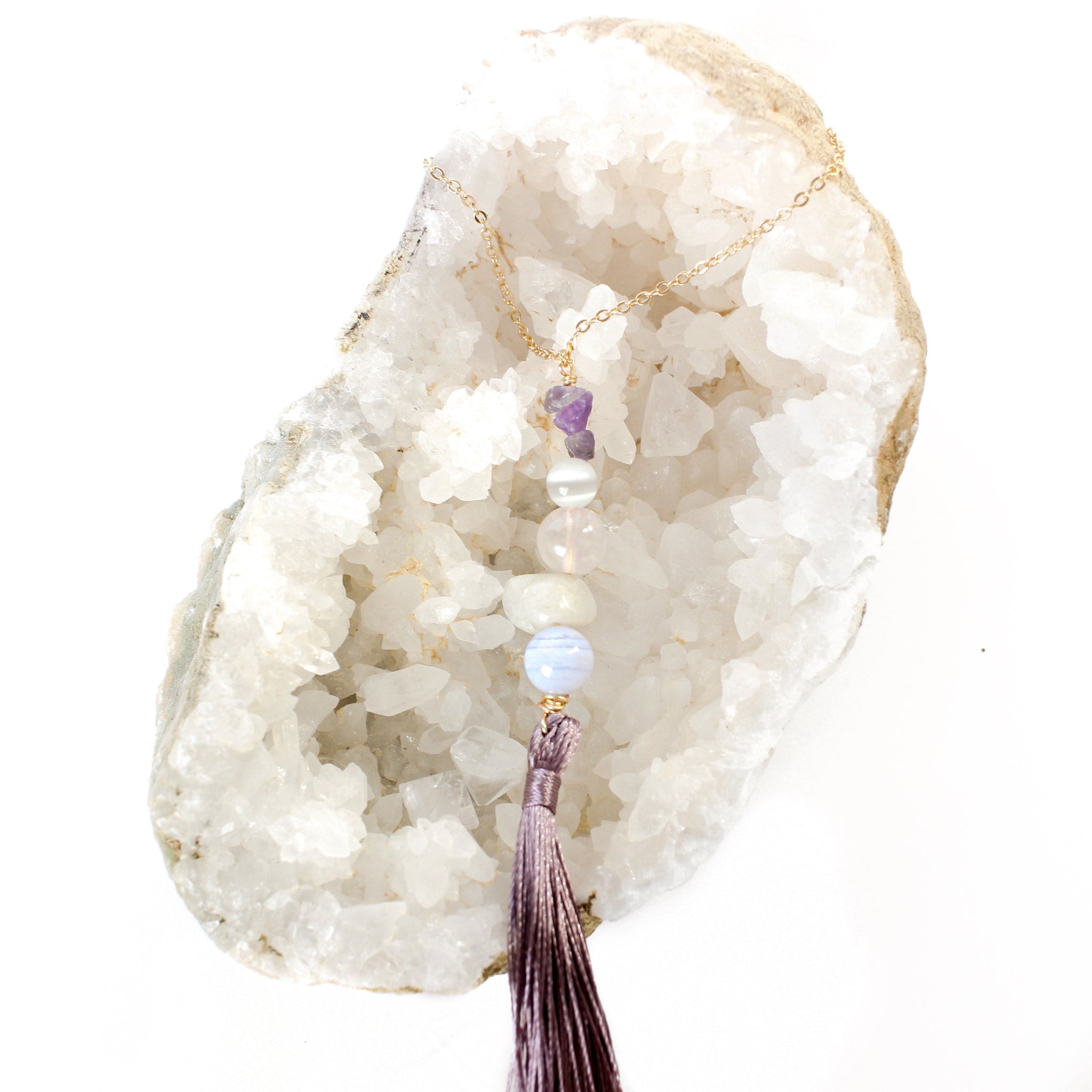 string of crystals with a loop of thin gold chain to hang it. the crystals from top to bottom are amethyst, cats eye, rose quartz, moonstone and blue lace agate. at the bottom to the crystals is a mauve-purple tassel. door tassel is laying on a bed of white crystals with a white background.