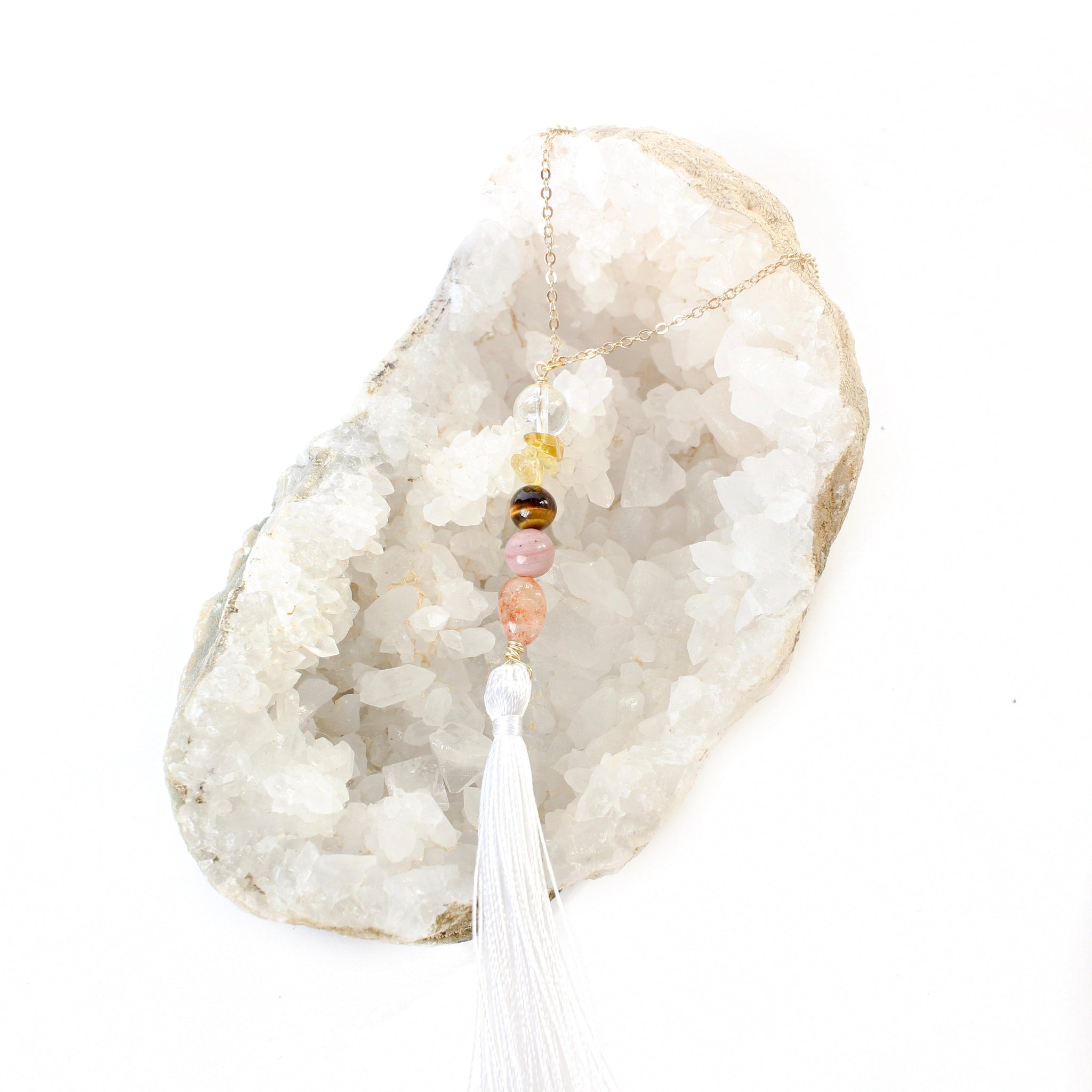 string of crystals with a loop of thin gold chain to hang it. the crystals from top to bottom are clear quartz, citrine, tigers eye, rhodonite and sunstone. at the bottom to the crystals is a white tassel. door tassel is laying on a bed of white crystals with a white background.