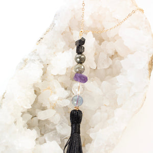 string of crystals with a loop of thin gold chain to hang it. the crystals from top to bottom are black tourmaline, pyrite, labradorite, amethyst, clear quartz and fluorite. at the bottom to the crystals is a black tassel.  door tassel is laying on a bed of white crystals with a white background.