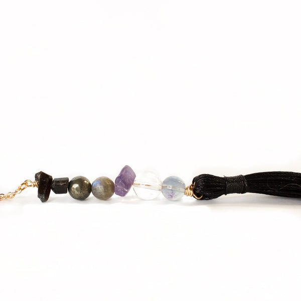 string of crystals with a loop of thin gold chain to hang it. the crystals from top to bottom are black tourmaline, pyrite, labradorite, amethyst, clear quartz and fluorite. at the bottom to the crystals is a black tassel.   white background.