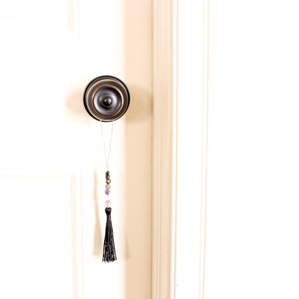 string of crystals with a loop of thin gold chain to hang it. the crystals from top to bottom are black tourmaline, pyrite, labradorite, amethyst, clear quartz and fluorite. at the bottom to the crystals is a black tassel.  door tassel is hanging on a black doorknob, on a white door.