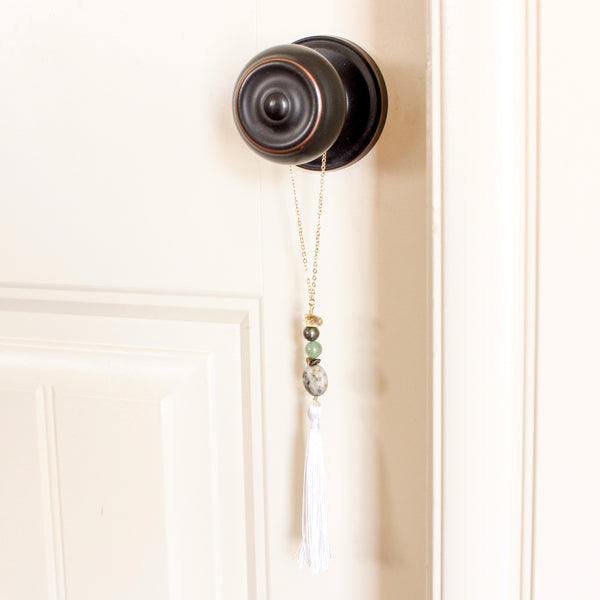 string of crystals with a loop of thin gold chain to hang it. the crystals from top to bottom are citrine, pyrite, green aventurine, tigers eye and pyrite in quartz. at the bottom to the crystals is a white tassel. door tassel is hanging on a black doorknob on a white door.