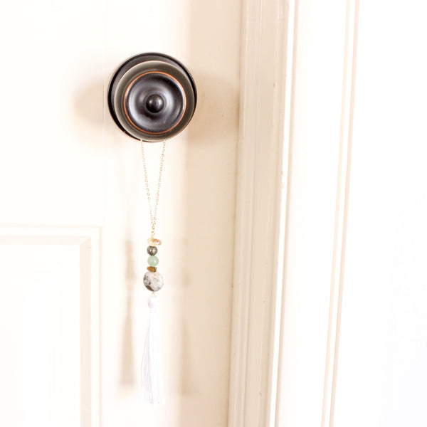 string of crystals with a loop of thin gold chain to hang it. the crystals from top to bottom are citrine, pyrite, green aventurine, tigers eye and pyrite in quartz. at the bottom to the crystals is a white tassel. door tassel is hanging on a black doorknob on a white door.