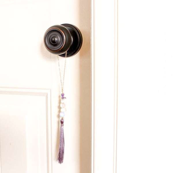 string of crystals with a loop of thin gold chain to hang it. the crystals from top to bottom are amethyst, cats eye, rose quartz, moonstone and blue lace agate. at the bottom to the crystals is a mauve-purple tassel. door tassel is hanging on a black doorknob on a white door.