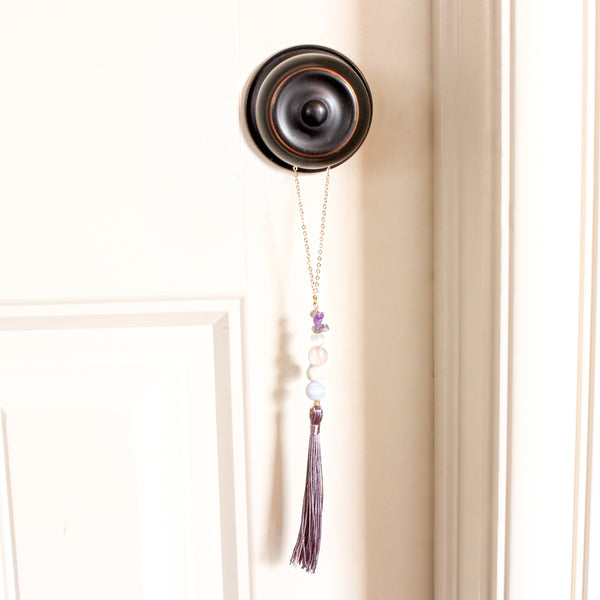 string of crystals with a loop of thin gold chain to hang it. the crystals from top to bottom are amethyst, cats eye, rose quartz, moonstone and blue lace agate. at the bottom to the crystals is a mauve-purple tassel. door tassel is hanging on a black doorknob on a white door.